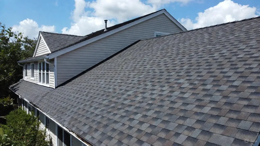 Close-up aerial view of a residential roof installed with dark gray, dimensional rubber roofing shingles under a bright, cloudy sky.