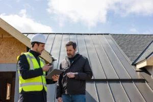 A roofing professional using a tablet to explain standing seam metal roof cost vs shingle pricing to a homeowner, with both a grey standing seam metal roof and traditional asphalt shingles visible in the background.